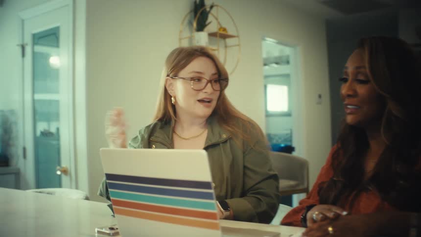 A woman with glasses explains a project on her laptop to a colleague as the camera slowly pushes in