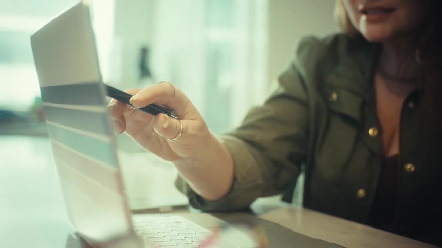 A businesswoman in a green jacket points to her laptop screen with a pen while presenting or discussing work