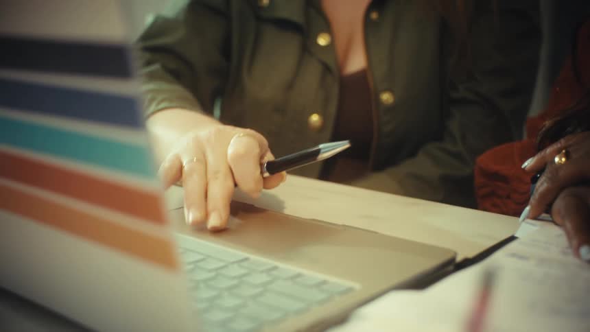 A close-up shot reveals a professional woman with glasses using her laptop to explain a project to a colleague during a meeting