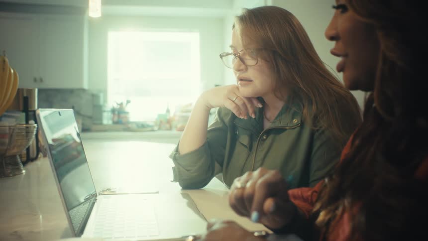 Two diverse women collaborate on a project using a laptop at a kitchen counter, pointing at the screen and discussing ideas