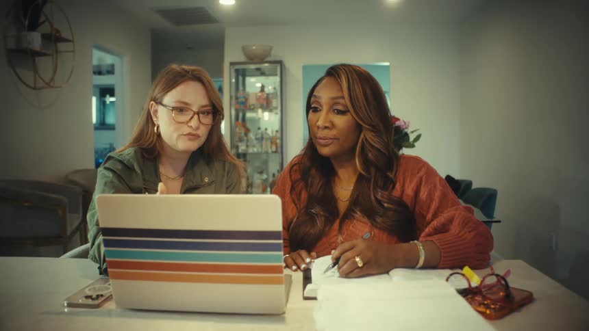 Two diverse women collaborate on a project from a home office, looking at a laptop and taking notes