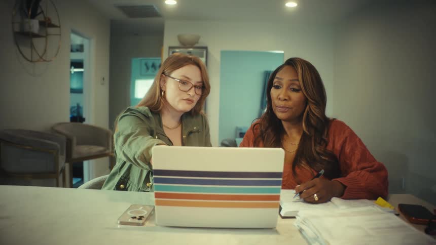 Zooming out from two diverse women collaborating over a laptop in a modern home office, one pointing at the screen and the other taking notes