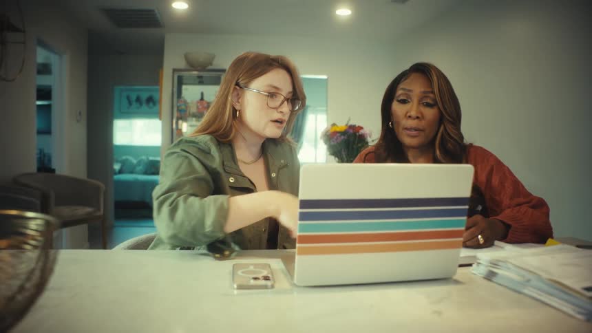 A young woman wearing glasses explains something on a laptop to an older woman as they sit together at a kitchen counter
