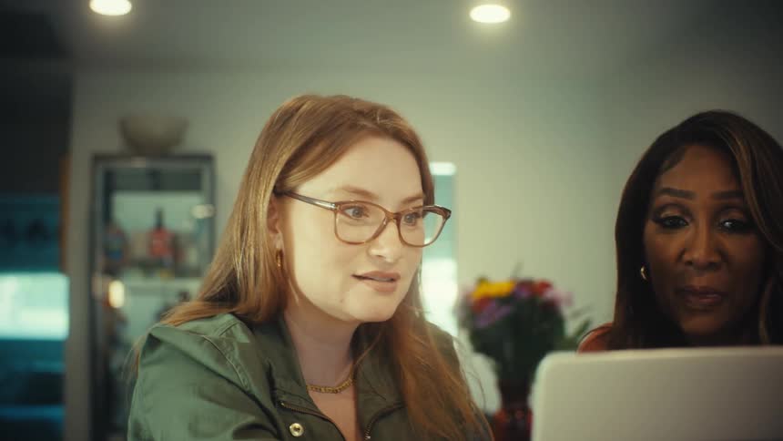 Two diverse women collaborate on a laptop in a modern home, their expressions shifting from happy to focused as they talk