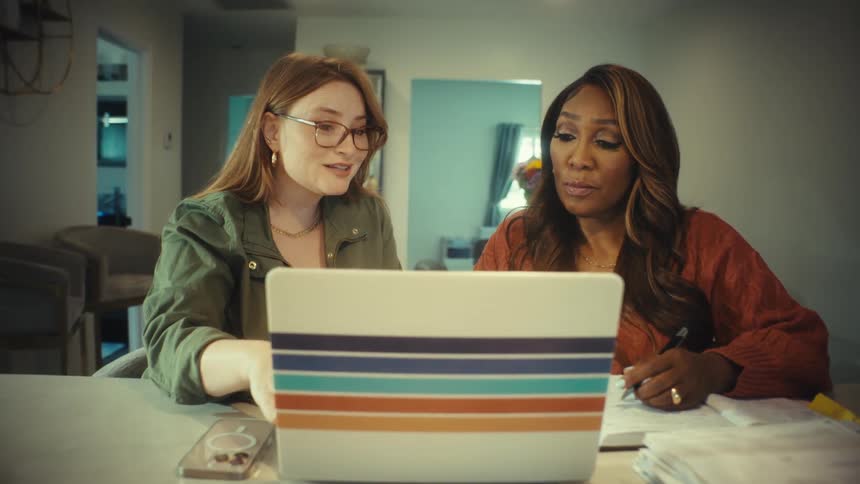 A slow push-in on two diverse women working together at a kitchen island, using a laptop and taking notes