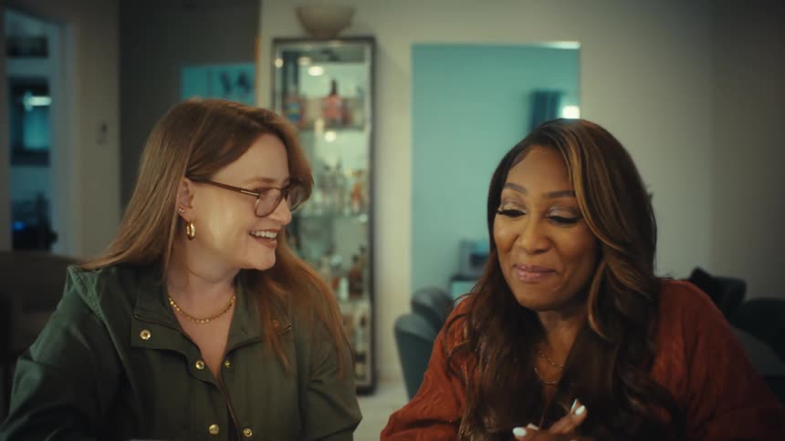 Two diverse female colleagues smile and shake hands while working together on a laptop in a modern home