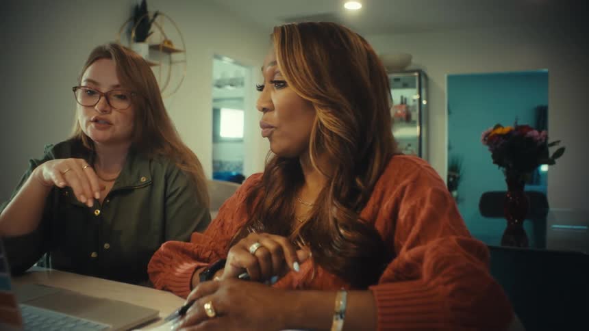 A slow push-in shot captures two diverse female colleagues collaborating on a laptop at a kitchen counter in a modern home