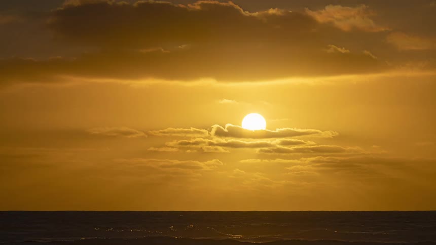 A stunning timelapse captures the golden sun rising over the ocean horizon through a layer of moving clouds