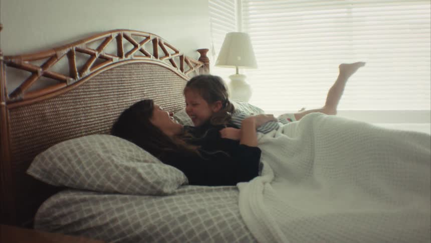 A joyful mother and her young daughter laugh and cuddle while playing together in bed on a bright morning