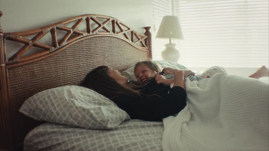A joyful mother and her young daughter laugh and cuddle while playing together in bed on a bright morning