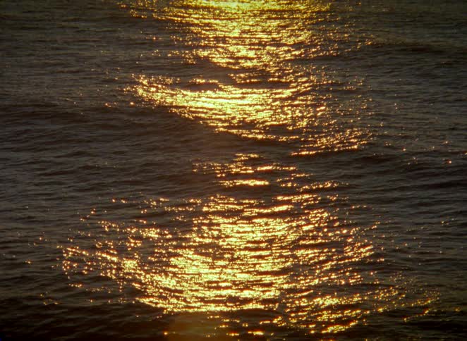 The tranquil ocean sparkles with golden light as the camera slowly reveals the sun setting over the horizon