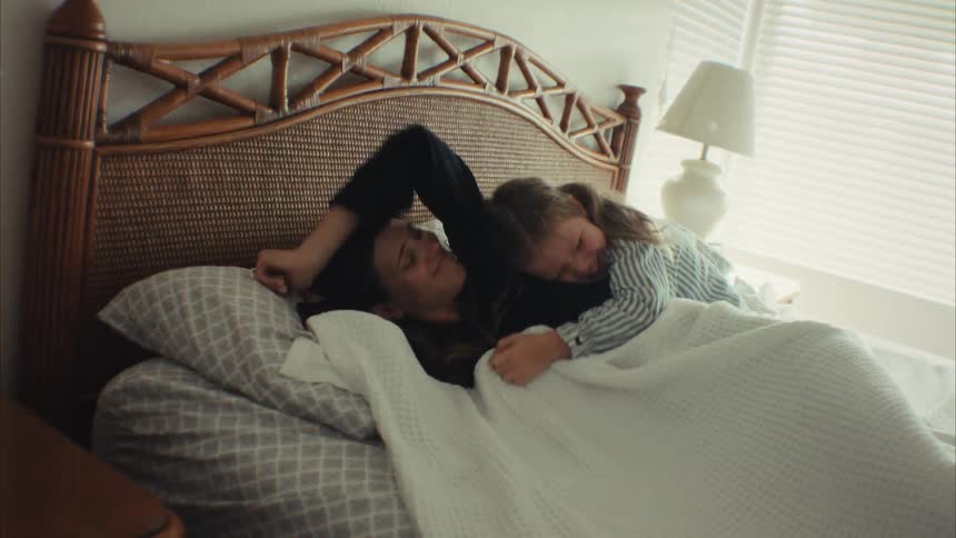 A playful young daughter crawls into bed and wakes up her sleeping mother by tickling her, and they share a warm morning cuddle