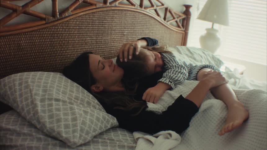 A playful young daughter crawls into bed and wakes up her sleeping mother by tickling her, and they share a warm morning cuddle