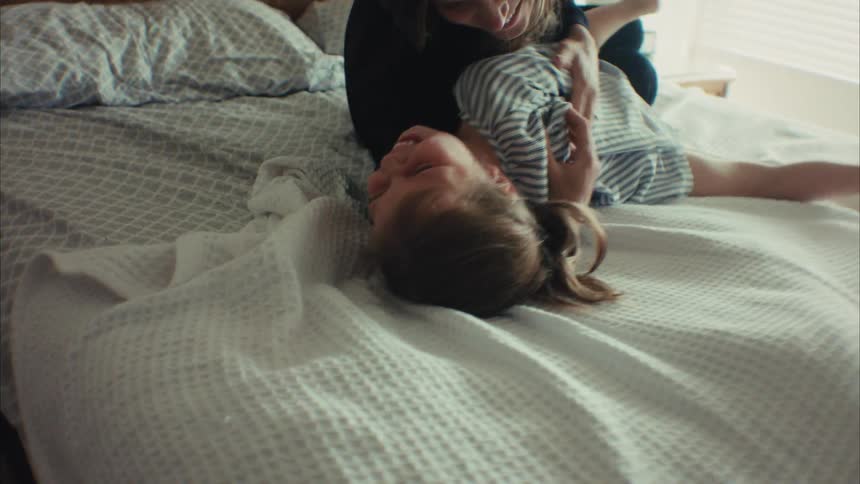 A joyful mother playfully tickles her laughing young daughter as they have fun together on a bed in a sunlit bedroom