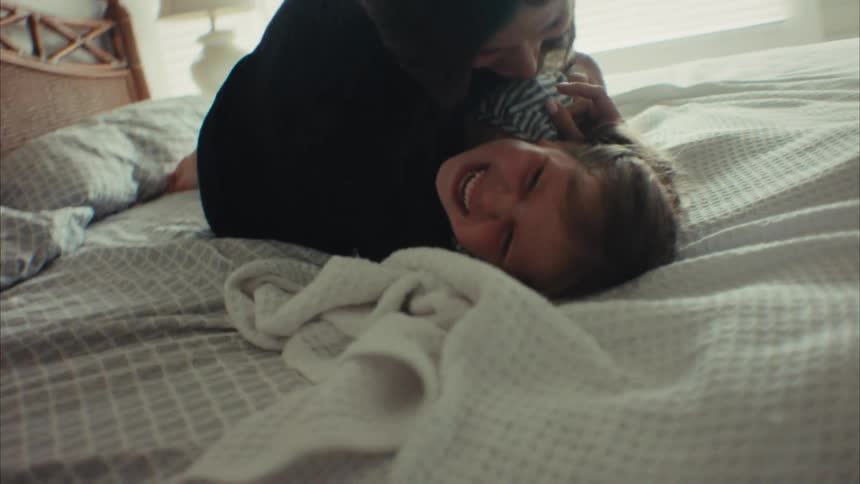 A joyful mother playfully tickles her laughing young daughter as they have fun together on a bed in a sunlit bedroom