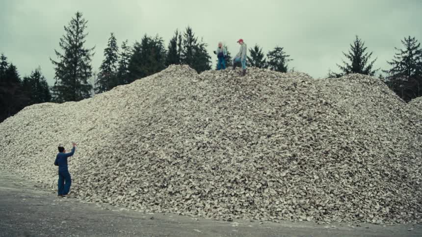 A group of people stand atop a massive mountain of discarded oyster shells at an oyster farm