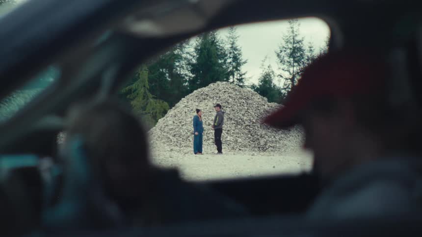 A man and woman talk in front of a large pile of oyster shells, as seen through the window of a truck