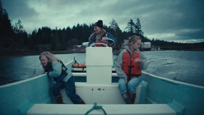 A mother steers a boat down a river in slow motion while her two happy daughters enjoy the ride on an overcast day