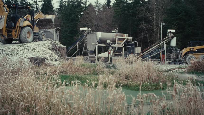 Heavy machinery operates at a rural rock quarry, processing gravel next to a murky pond