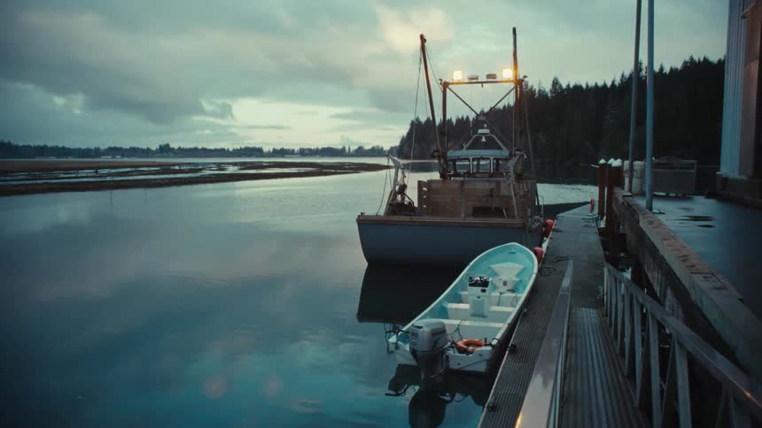 A fishing boat and a motorboat are moored at a quiet dock on a calm river under a moody, overcast sky at dusk