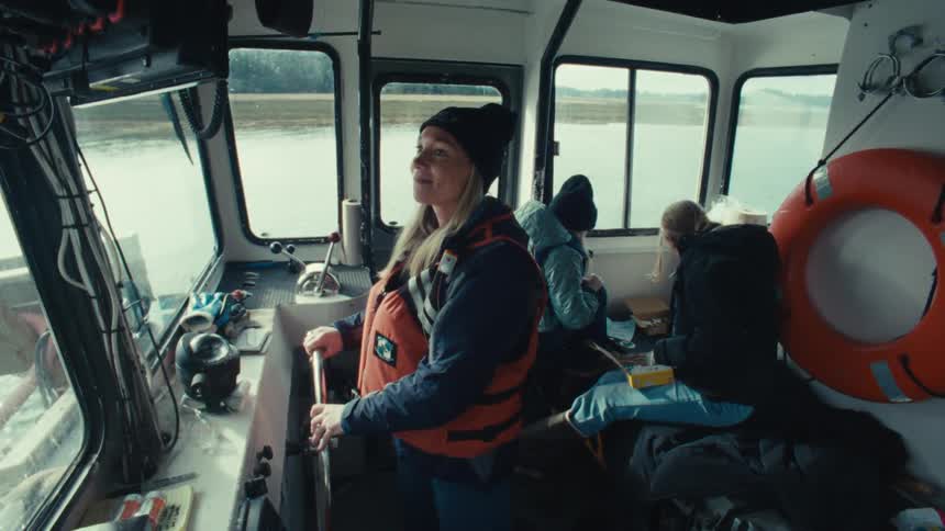 A female boat captain confidently steers her vessel through calm waters on an overcast day