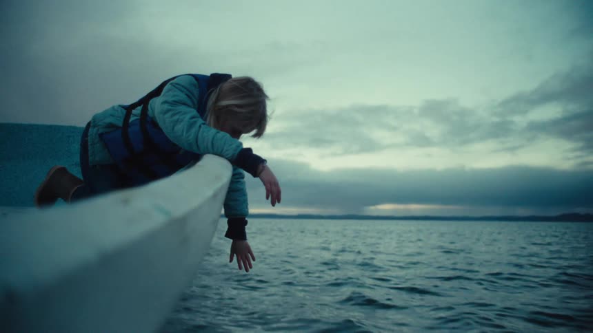 Slow-motion shot of a curious child in a life vest reaching for the water from the bow of a boat on a calm, overcast day
