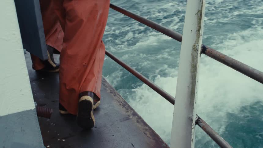 Close-up on a fisherman's boots as they stand on the edge of a boat with the ocean wake rushing by