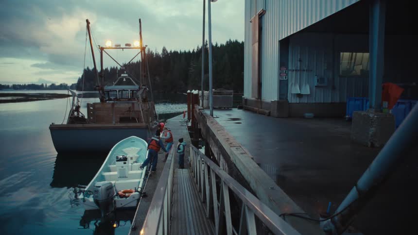 A group of people disembark from a small boat at a commercial fishing dock on a calm, overcast morning