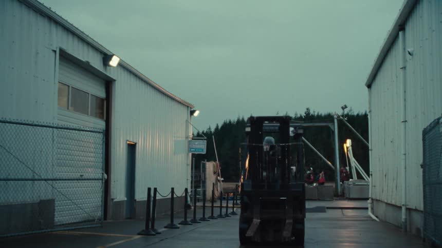 A forklift operator carefully backs up the vehicle between two large industrial warehouses during the early morning hours