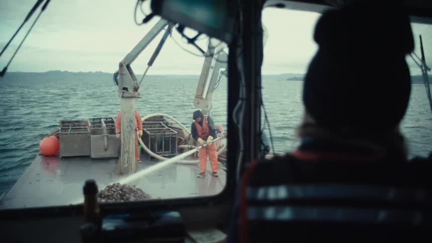 View from inside a fishing boat cabin as a deckhand sprays down a fresh catch of oysters on a gray day at sea