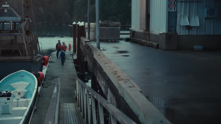 A family in life jackets walks down a wet wooden dock at a fishing marina during a cool, overcast day