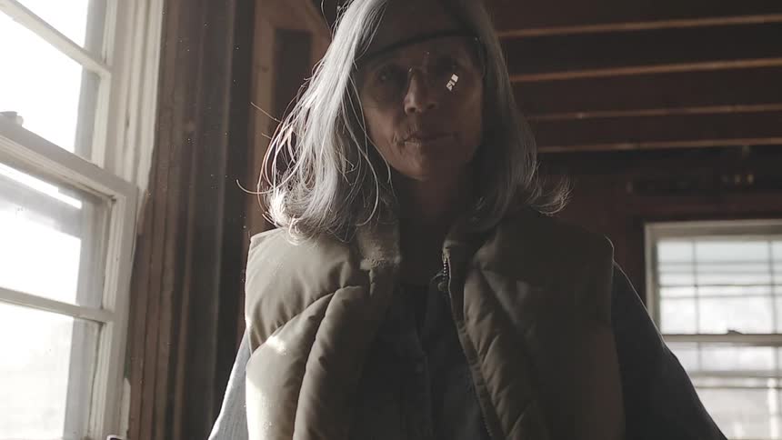 A determined senior woman holding a sledgehammer looks directly at the camera in a dusty, backlit room under demolition