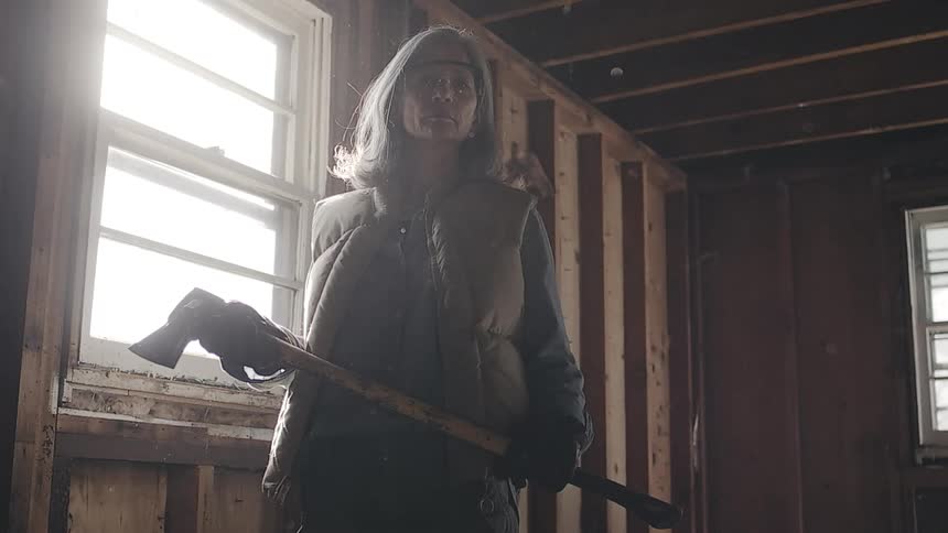 A determined woman stands with an axe in a dusty room under demolition as particles float through the air in slow motion