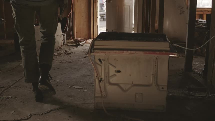 A dramatic slow-motion dolly shot reveals a person in work boots walking through the dusty debris of a demolished house while holding a large maul