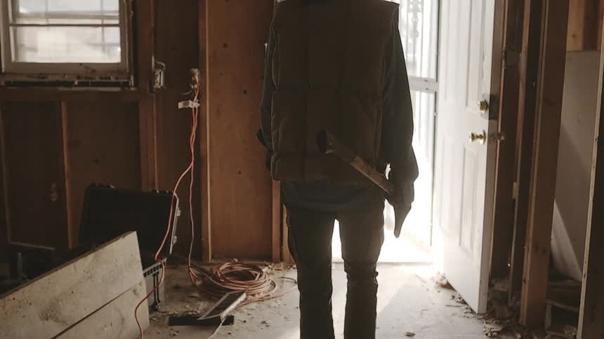 A person in boots walks in slow motion with an axe through a demolished and gutted house toward a bright doorway