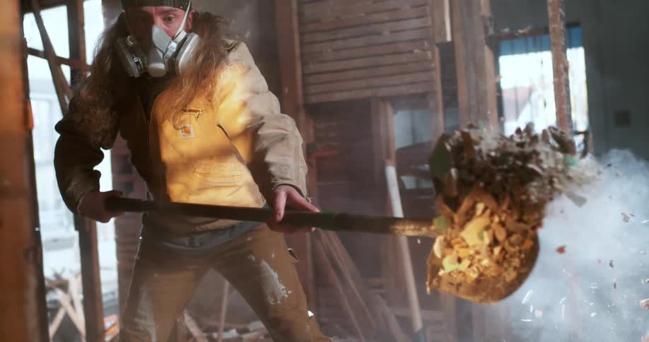 A demolition worker wearing a respirator mask violently throws a shovelful of drywall and debris towards the camera in ultra slow motion