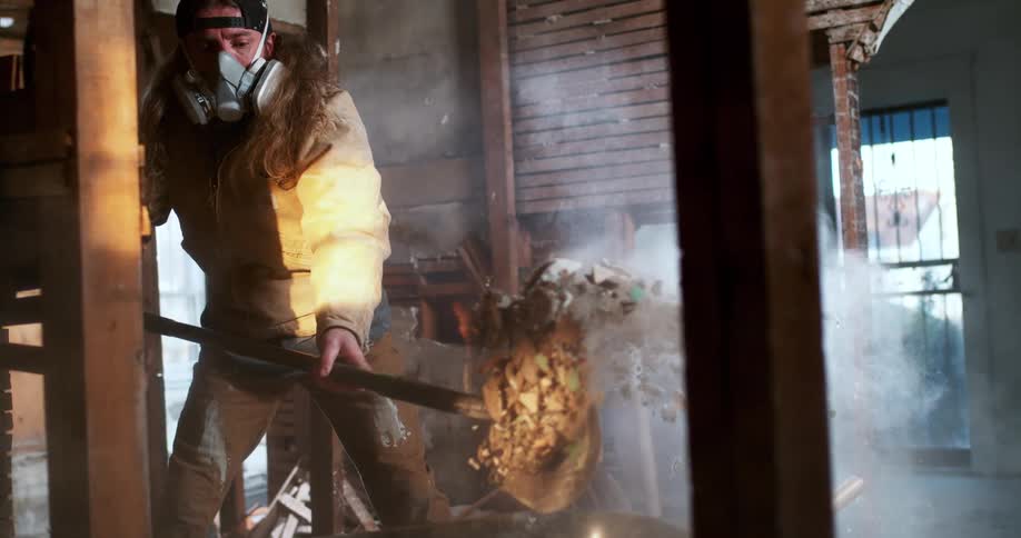 A construction worker wearing a respirator mask shovels a load of dusty debris into a wheelbarrow in dramatic slow motion