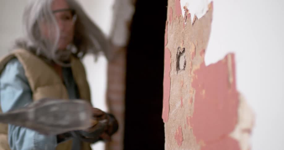 A woman in protective gear smashes a hole through a dilapidated interior wall with a sledgehammer in slow motion