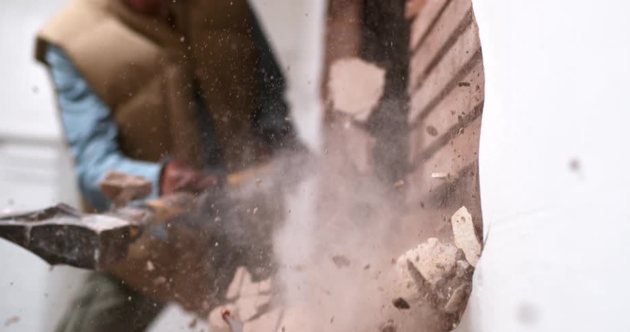 A worker demolishes an interior wall with a pickaxe, sending debris flying in dramatic slow motion