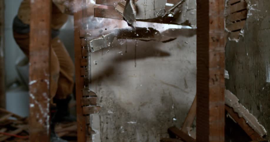 Dramatic slow-motion footage shows a plaster and lath wall being demolished, shattering towards the camera