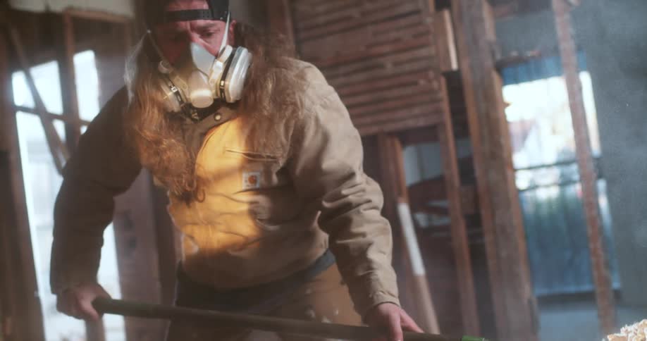 A construction worker in a respirator mask shovels dusty debris into a wheelbarrow at a demolition site in slow motion