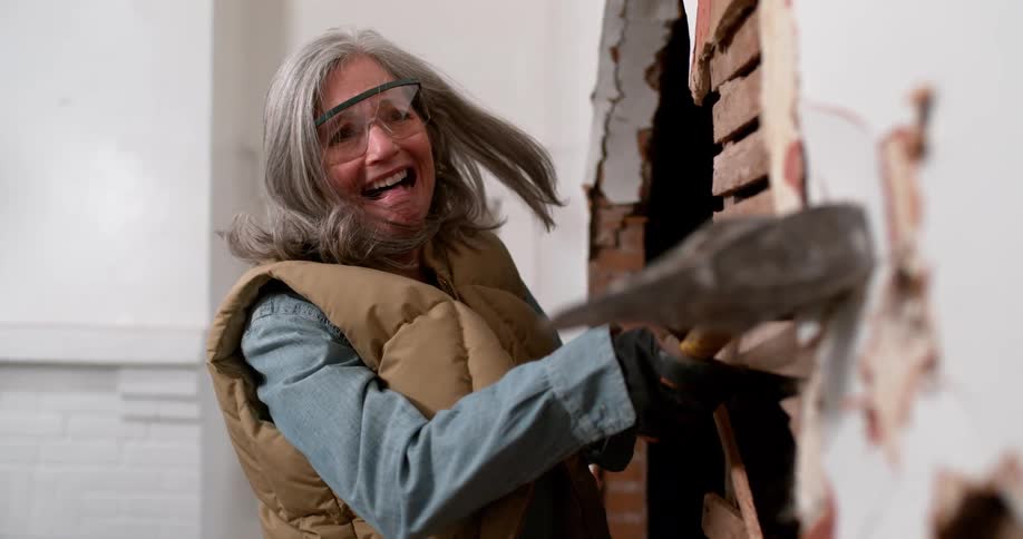 Empowered older woman joyfully demolishes an interior wall with a sledgehammer in exhilarating slow motion