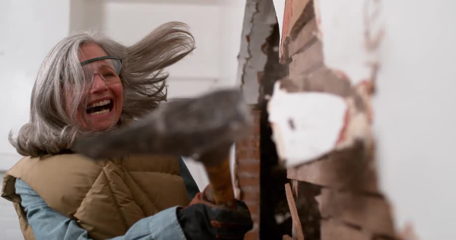 An empowered senior woman laughs with joy while demolishing an old plaster wall with a sledgehammer in slow motion
