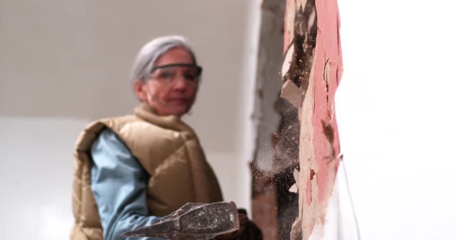 A determined older woman in safety glasses smashes an interior plaster wall with a heavy hammer in super slow motion