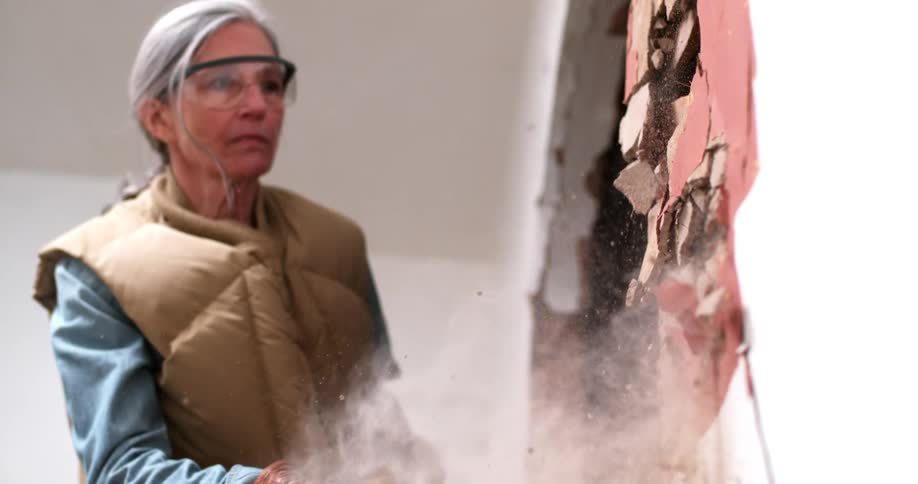 A determined woman wearing safety glasses demolishes an interior wall with a sledgehammer, sending plaster dust flying in super slow motion