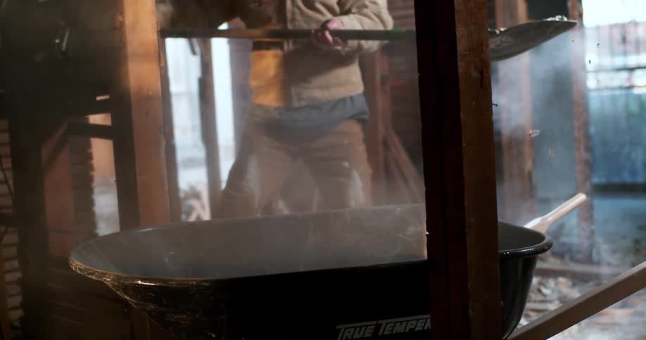 A construction worker wearing a respirator mask shovels dusty debris into a wheelbarrow in a dilapidated building in dramatic slow motion