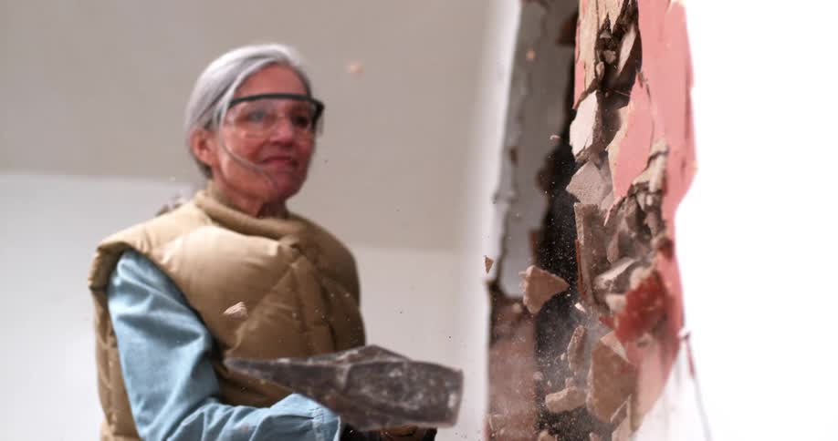 Super slow motion low angle shot of an older woman smashing a wall with a sledgehammer during a home renovation