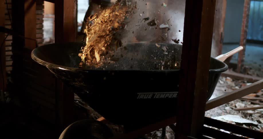A construction worker in a respirator mask shovels dusty demolition debris into a wheelbarrow in dramatic slow motion