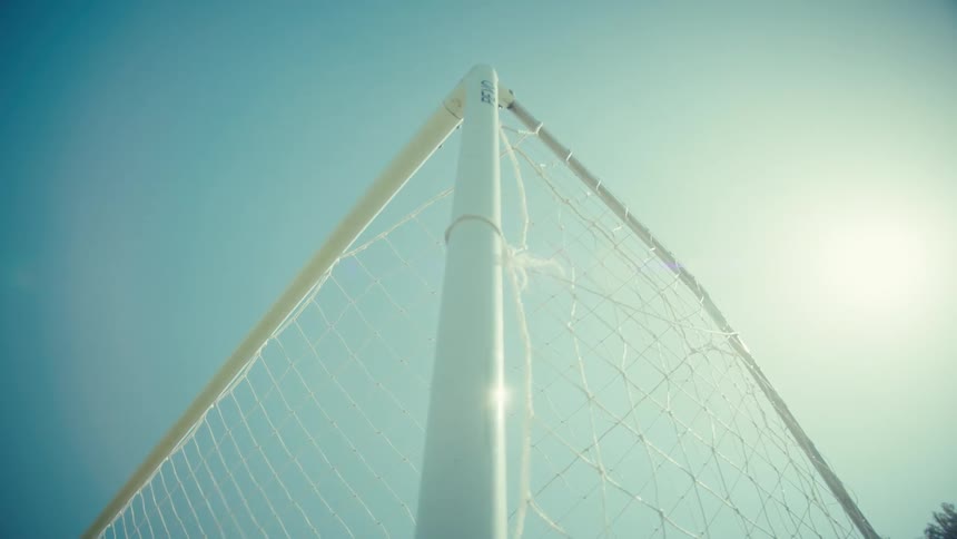 A soccer ball is kicked into the top corner of a goal in slow motion against a bright blue sky