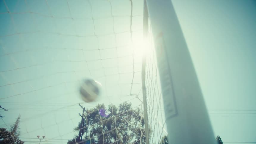 A soccer ball scores a goal as it hits the back of the net in slow motion on a sunny day